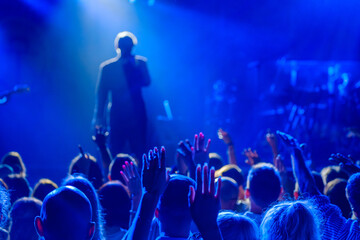 Back view of crowd of unrecognizable people dancing with raised arms while standing near stage during concert of male singer and musician in club