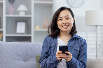 Fototapeta na wymiar Portrait of a young beautiful Asian woman at home on the couch, woman smiling and looking at the camera holding a telephone, smartphone application user, satisfied and happy.