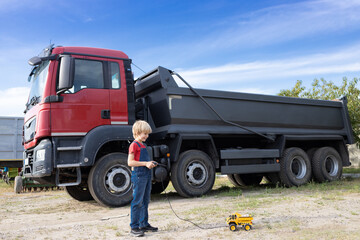 boy is playing with toy dump truck. Behind child is real large truck. boy's hobby for construction equipment. Be a driver or a mechanic like dad. commercial vehicles for business on credit or rent