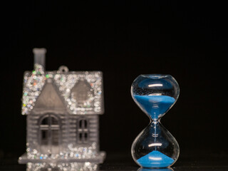 Sand clock with blue sand in focus. Glamourous house with glitter out of focus in the background. Dark background. Time to buy house concept. Invest in real estate theme. Banking and finance industry