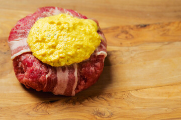 Big uncooked beef burger wrapped with bacon slice and portion of cheese on wooden board. Premium meat product. High quality cheeseburger.