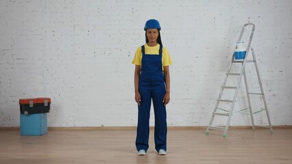 Full length shot of a smiling dark-skinned young female construction worker in a uniform and hardhat standing in the room under renovation.
