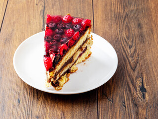 Slice of tasty multi layered cake with raspberry and blue berry in jelly on top. Portion of delicious dessert on white plate and wooden table. High quality bakery product serving in coffee shop.