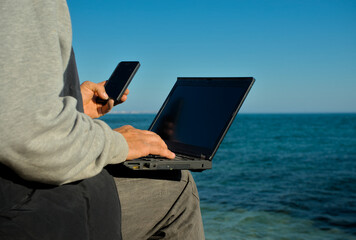 A man with a laptop and phone in his hand sits on the seashore and works. Remote remote work from anywhere in the world