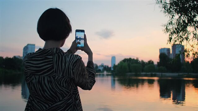 Woman takes photo of lake with mobile phone. Beautiful girl takes pictures and videos on mobile phone of a sunset on summer evening