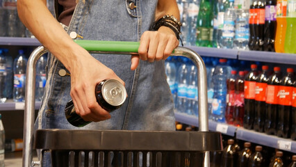 Close-up of a young woman's hands putting a can of soda or enerdy drink into a shopping cart