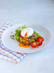 Runny poached egg on wholemeal bread with salad, tomato, red onion, lettuce and cucumber. Decorated with cherry tomatoes, microgreens, sesame and pumpkin seeds on white plate on cotton chessboard