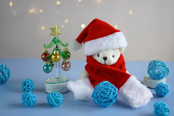 a white teddy bear in a red hat and Santa Claus scarf sits near a glass elegant Christmas tree with decorations in the form of colorful balls on a table on a gray background with garlands of bokeh