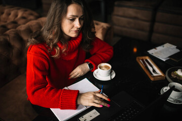 woman in a red sweater works in a coworking cafe with a laptop and a notebook, freelance work in winter, work on the Internet