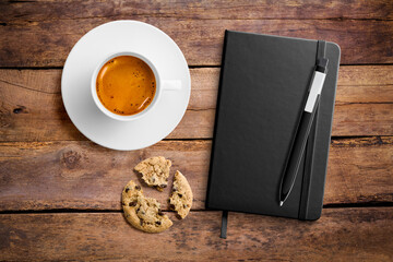 Top view of a blank hardcover leather notebook on a wood office desk table with a cup of coffee and a chocolate cookie. High resolution.
