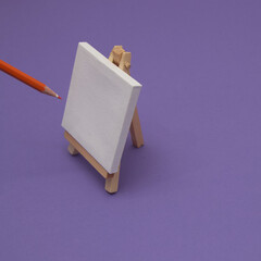 Painting canvas on a stand on a purple background with an orange crayon. Minimal painting composition.