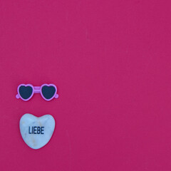 A heart-shaped stone that says love in German, and sunglasses in the lower left corner on a red background. Minimalistic flat lay with summer motifs.