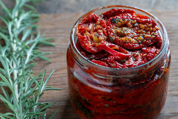 Sun-dried red tomatoes with garlic, green rosemary, olive oil and spices in a glass jar on a wooden table. Rustic style, closeup