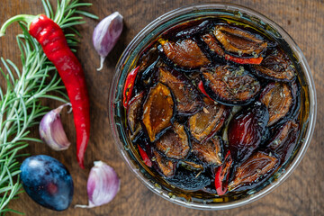 Sun-dried plums with garlic, green rosemary, red chili pepper, olive oil and spices in a glass jar on a wooden table. Rustic style, top view, close up