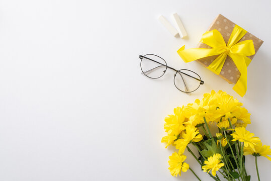 Celebrate Teacher's Day uniquely. A top-view picture showcases chalk, glasses, bunch of chrysanthemums and a gift box on a white isolated backdrop. Ample space for text or promotional content