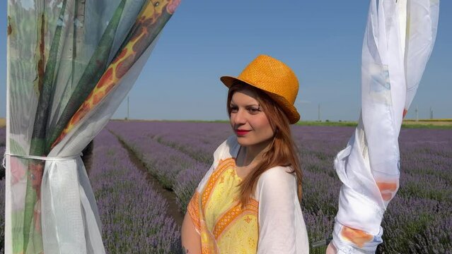 Pull out from close-up of young woman to medium shot of decorative door in lavender field. Woman fanning herself with orange hat. Photo session. Natural woman in hippie style. Model. Actress.