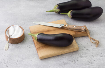 Fresh eggplant on a wooden board, a bowl of salt and a knife on a gray textured background. Cooking delicious vegan food