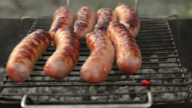Grilled sausages. Cook man preparing grilling food bbq. Barbecue with smoke, flame outdoors. Tasty juicy german bratwurst. Charcoal kettle grill outside in backyard. Family summer vacation. Close-up	

