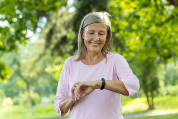 Senior beautiful gray-haired woman running in park, pensioner resting and using fitness bracelet of...