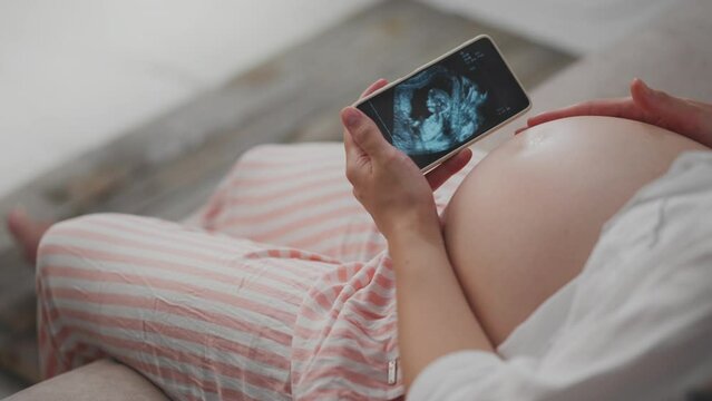 Mom is waiting, cute mom in comfortable pregnant clothes is holding phone in her hands on the screen, shot of the baby's reins. Woman examining the reins of her child holding a smartphone in her hands