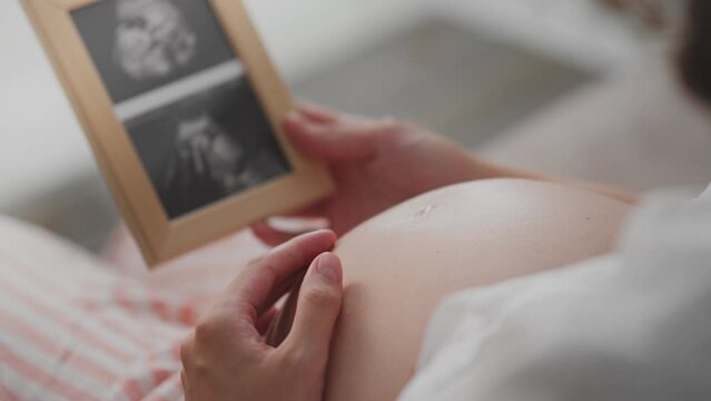 Pregnant, cute woman in light clothes is looking at picture of her unborn child's bridles on home bed. Camera movement modern woman holding a frame with a photo of the bridle baby care methods, birth