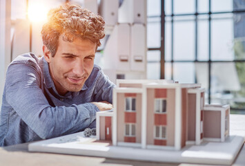 Young Male Designer Assembling Wooden Pieces into a House Miniature on Top of the Table with Blueprint.