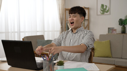 excited Taiwanese male employee working from home is screaming with clenched fist for getting pay raise online notification on the laptop in the living room
