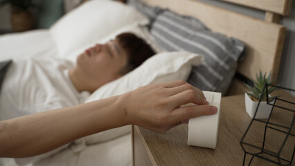 selective focus of a tired asian male’s hand reaching out for the ringing alarm clock on nightstand and stopping it to sleep longer on bed in the morning at home