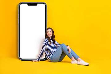 Full body photo of positive nice lady sit floor look huge empty space telephone screen isolated on yellow color background