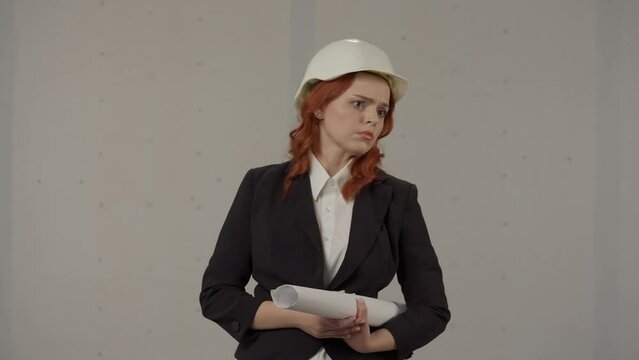 A woman architect indignantly points at someone, swears and screams. Portrait of a woman in a protective helmet and with blueprints in the studio on a gray background.