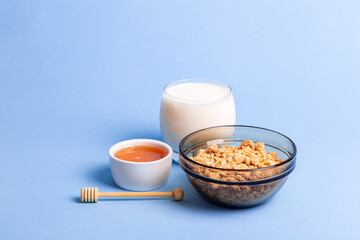 Glass bowl of granola, glass of milk and honey on blue background. Healthy and tasty breakfast.