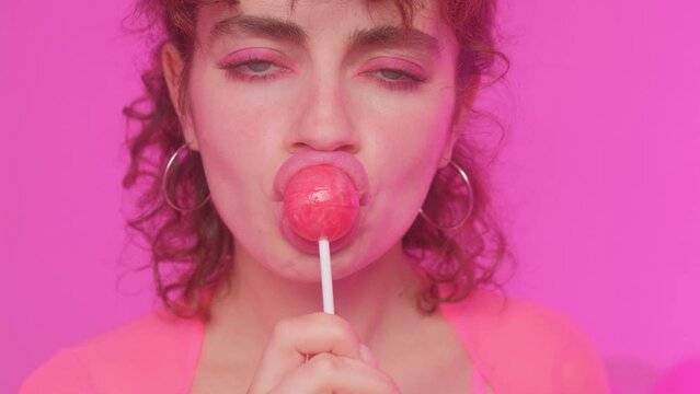 Glamorous beautiful young woman model on a pink background, eating a fudge lollipop. Girl licks a candy.