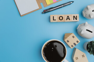 Top view, flat lay a coffee in cup, little homes, note book, 2 white piggybanks, and text LOAN on cubes on the blue desk office Financial, Accounting, Banking, Investment of real estate loan concept.