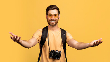 Happy middle aged man in sunglasses outstretching hands to camera, standing with backpack, yellow background