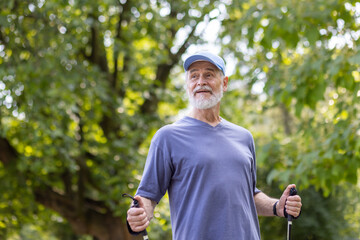 Senior gray-haired pensioner man on a walk in the park uses walking sticks, happy grandfather...