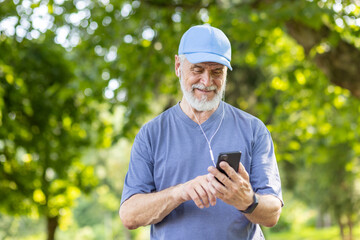 Senior gray-haired male athlete jogging in the park wearing headphones resting and listening to...