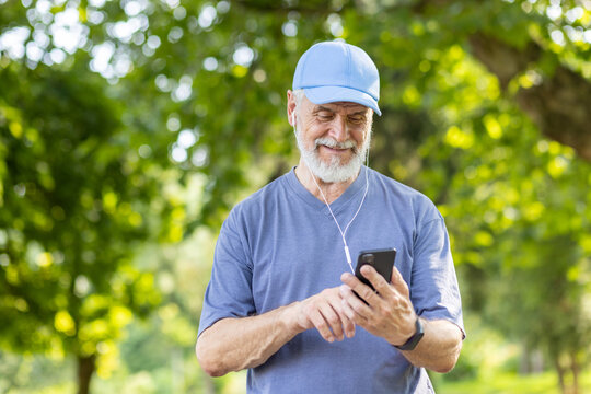 Senior gray-haired male athlete jogging in the park wearing headphones resting and listening to music online, using smartphone app, pensioner leading an active lifestyle.