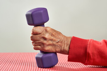 Wrinkled hand of elderly senior person holding violet small dumbbell on a red gym mat....