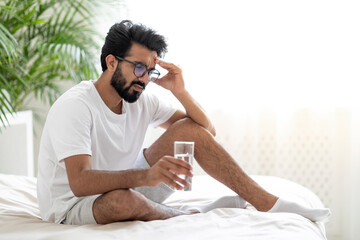 Hangover. Sick Indian Man Holding Glass Of Water While Sitting In Bed
