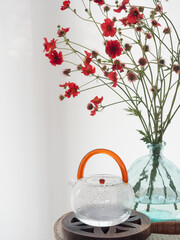Transparent glass teapot with orange handle with hot water on a small wooden table next to a vase with orange flowers. Simple decoration, harmony at home.