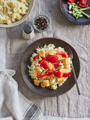 dish with rice, red bell pepper, spring onion and chicken on dark ceramic plate with fork, salt and pepper on the table. View from above.