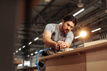 Young carpenter sanding wood piece in workshop in furniture factory