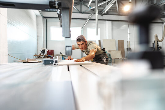 Young carpenter cutting a piece of wood in using a circular saw in furniture factory