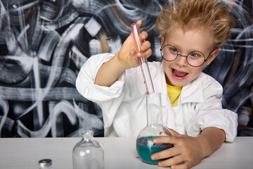 A young smart boy in round glasses and a white coat conducts chemical experiments in a real...