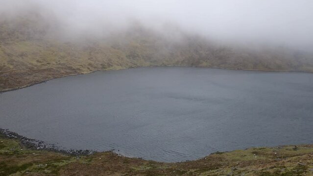 On a blustery day in Ireland, the surface of a serene highland lake is adorned with the ethereal presence of low-flying clouds, painting a captivating tableau.