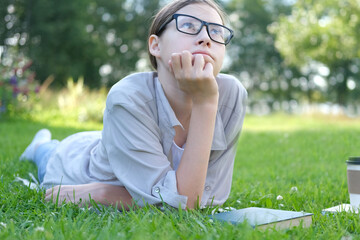 Teenage schoolgirl studying reading her books, lying down outdoors. Back to school, dreaming, inspiration. Student girl reads book on the green grass in the college yard or park. Distance learning.