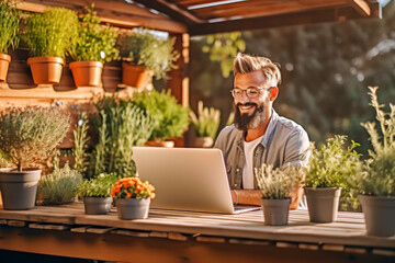 Happy hipster businessman florist in apron sitting with laptop at work in small sustainable floral shop. Small business owner works in flower shop and customer service . Startup. Own business concept