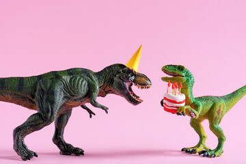 Cute happy green dinosaurs in birthday hats holding cake with flaming candles on pastel pink background. Copy space. Minimal art birthday card idea.