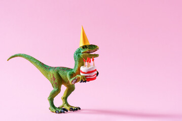 Cute happy green dinosaur in birthday hat holding cake with flaming candles on pastel pink background. Copy space. Minimal art birthday card idea.