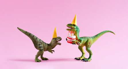 Fototapeta premium Cute happy green dinosaurs in birthday hats holding cake with flaming candles on pastel pink background. Copy space. Minimal art birthday card idea.